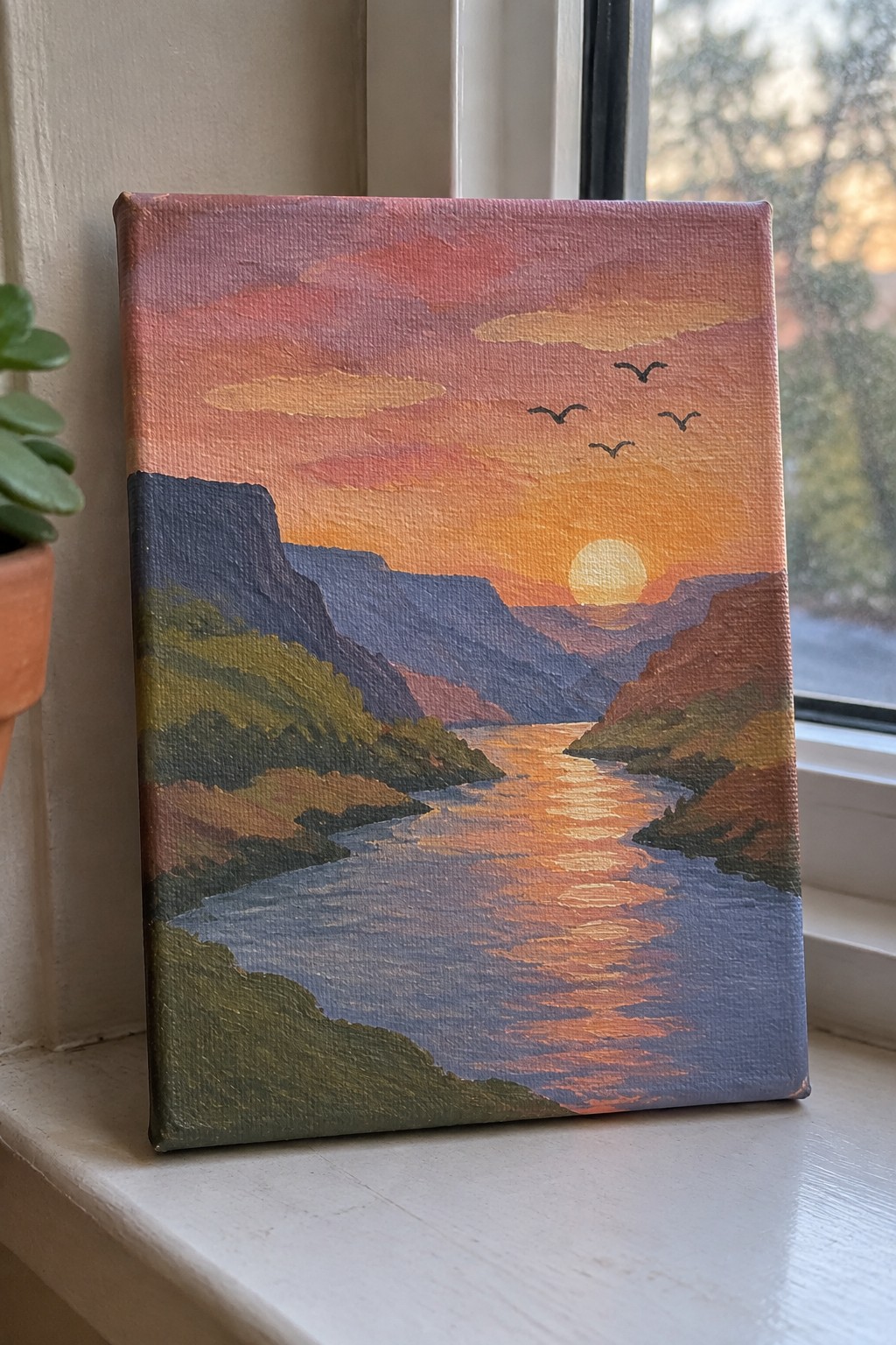 Sunset river valley painting with mountains and birds on windowsill beside potted plant.