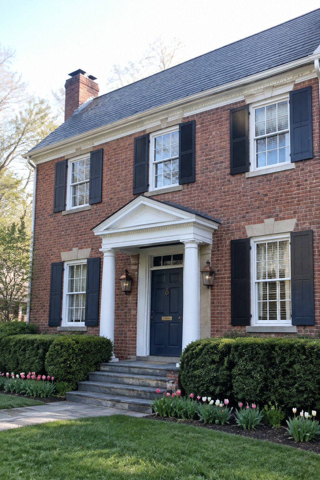 Two-story red brick house with black shutters, blue door, white columns, and tulip garden.