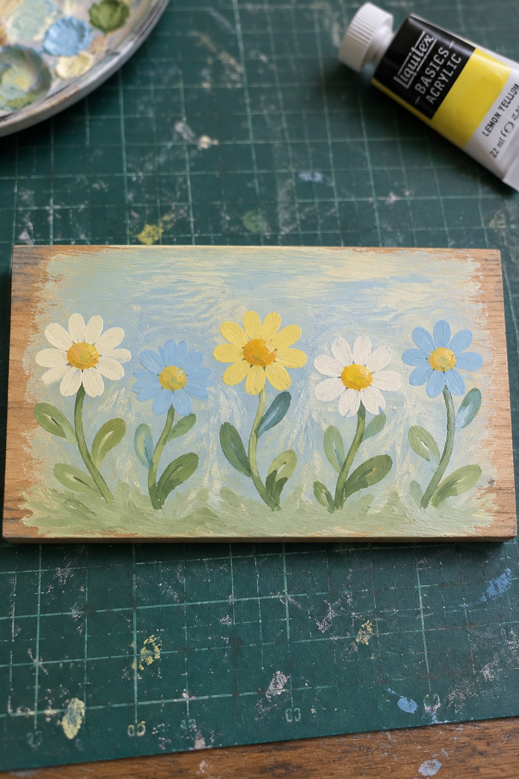 Hand-painted wooden plaque with white and blue daisies beside yellow acrylic paint tube