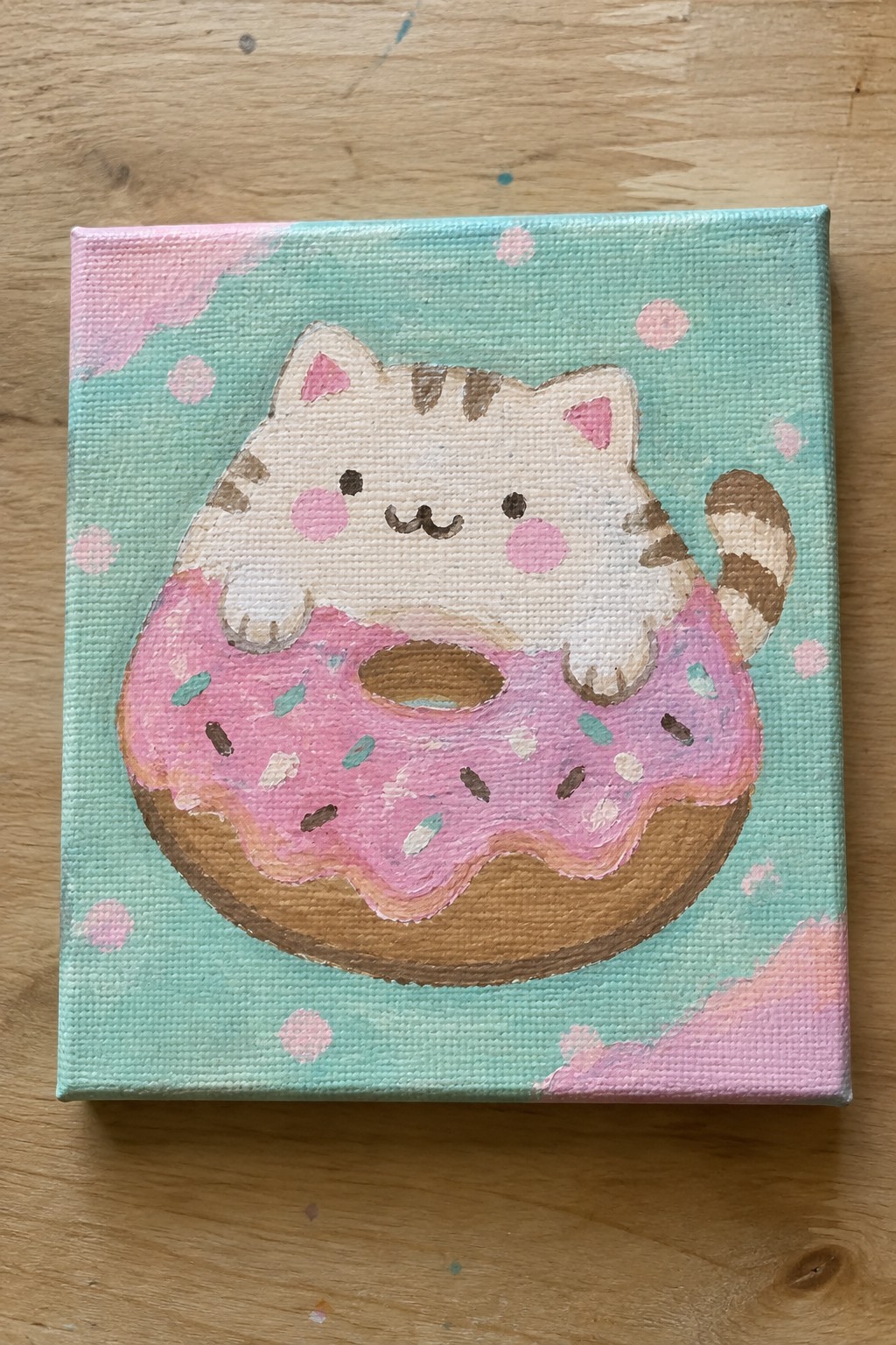 A small square canvas shows a white and brown striped cat with pink cheeks sitting on a pink frosted donut against a mint background with pink dots.