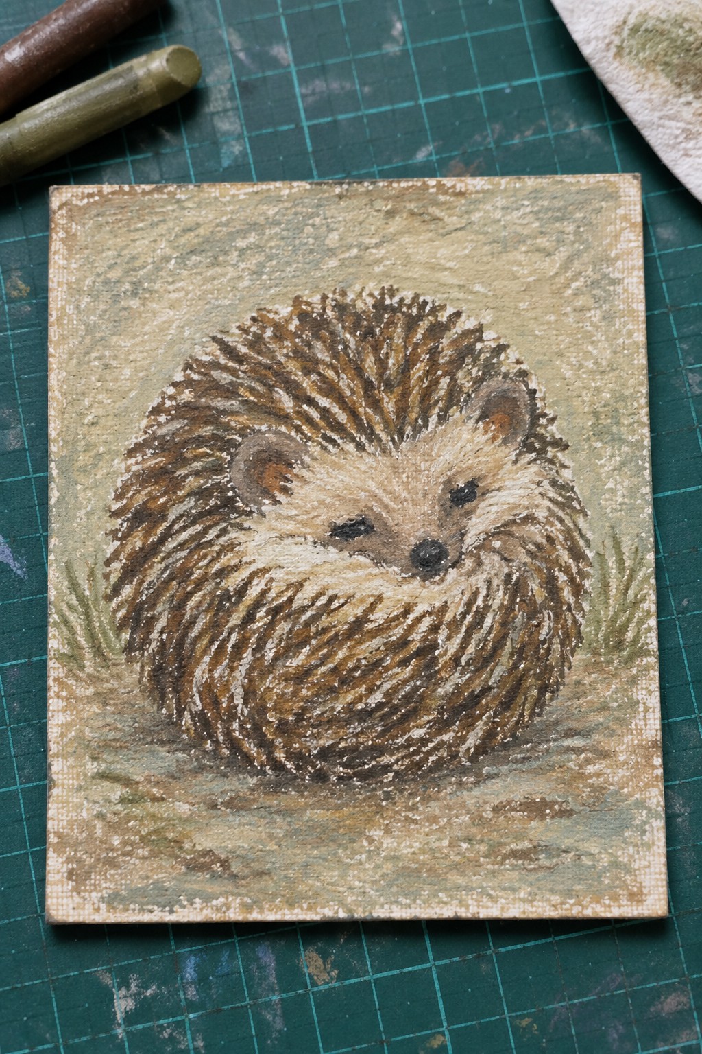 Pastel painting of curled hedgehog on small canvas over green cutting mat