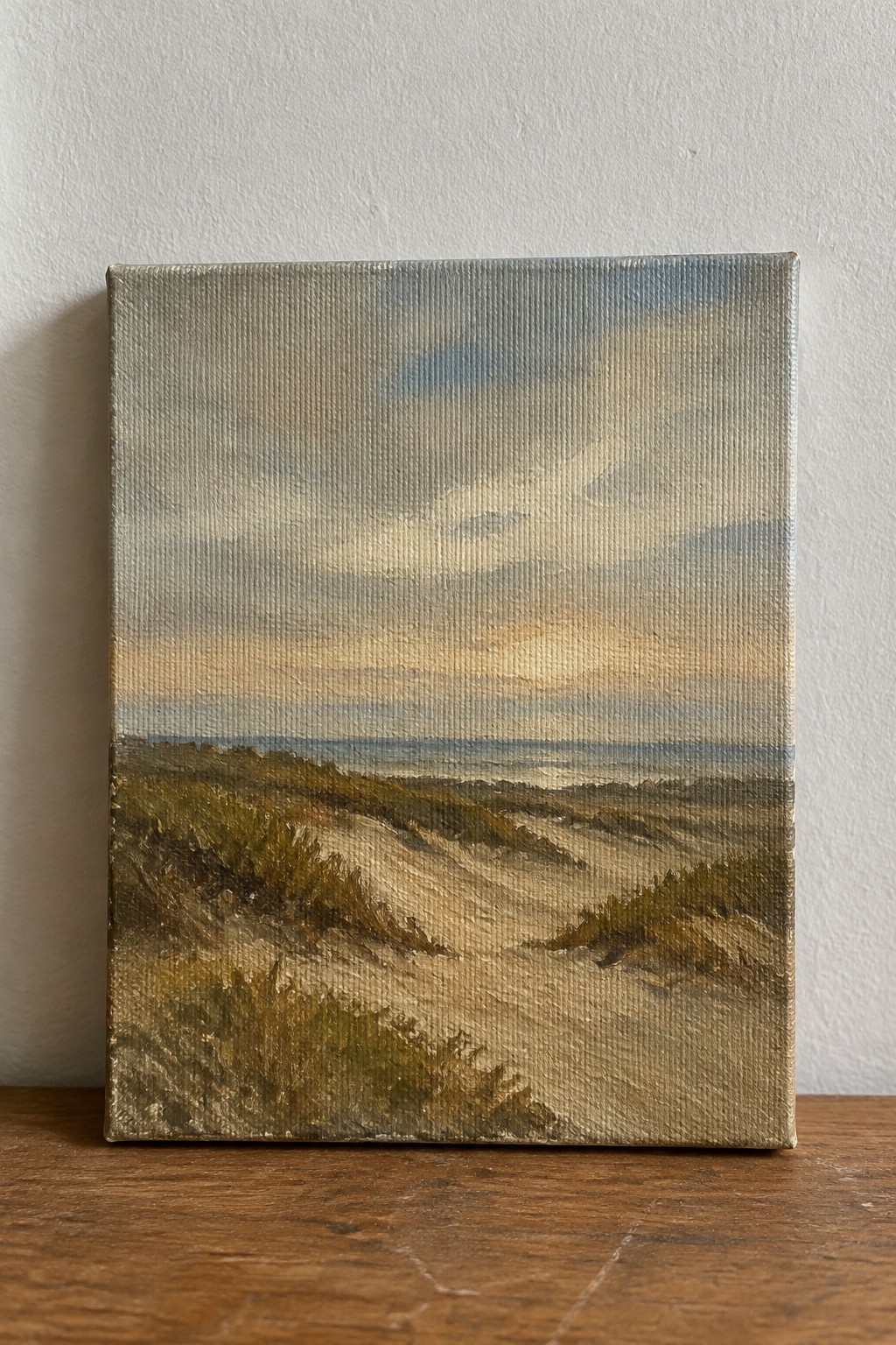 A small canvas painting of sand dunes with grass patches under a cloudy sky with warm light, resting on a wooden surface against a white wall.