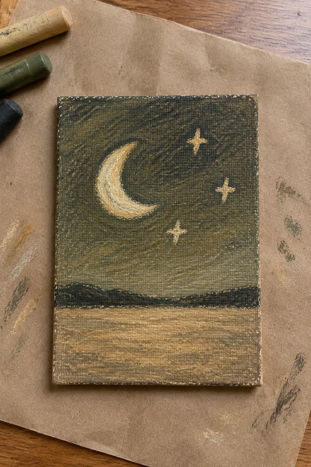 Miniature nightscape painting with golden crescent moon, stars, and dark landscape beside oil pastels.