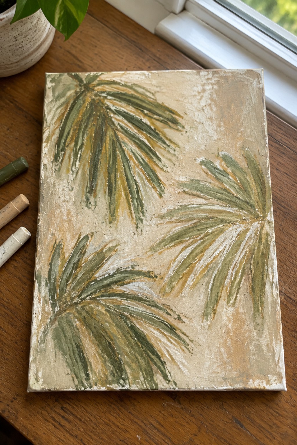 Tropical palm frond painting on canvas with pastels and plant on wooden table.