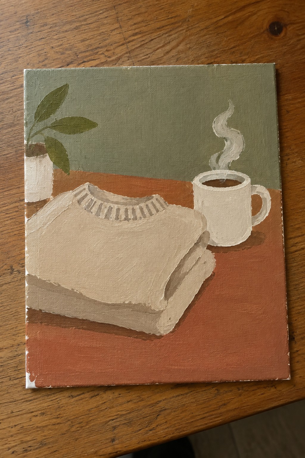 Folded beige sweater beside steaming white mug and potted plant on wooden table