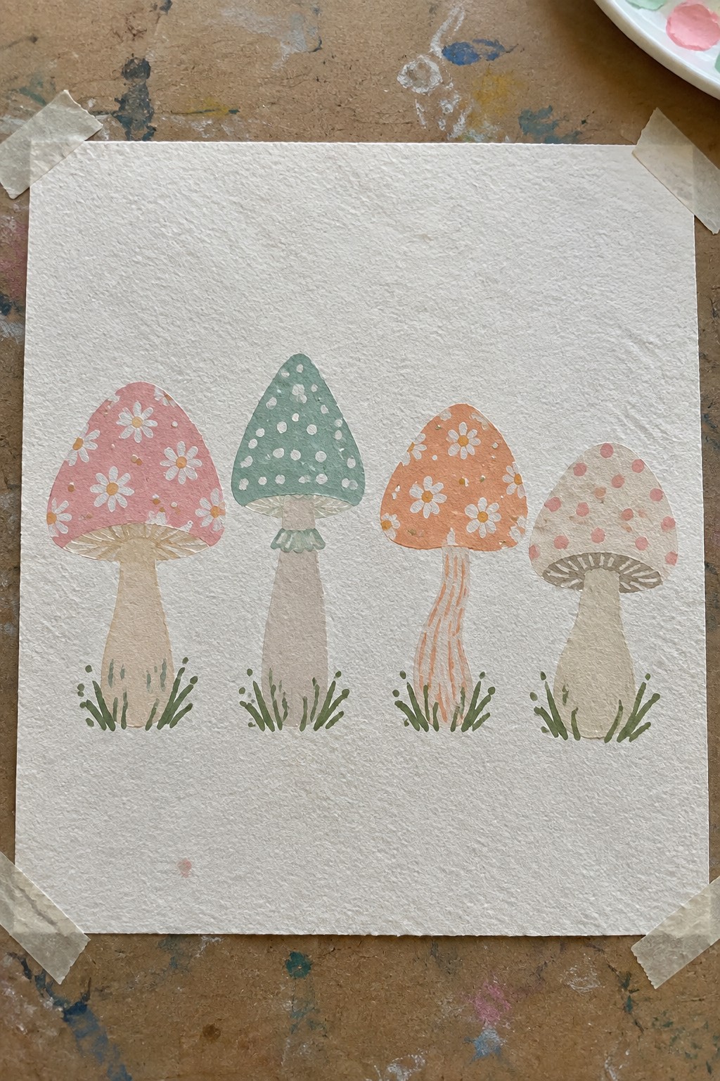 Four whimsical watercolor mushrooms with floral and dotted caps on taped paper.