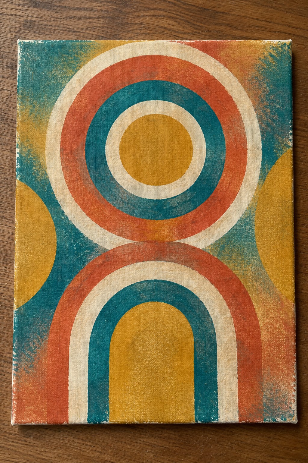 Abstract canvas with teal orange yellow concentric circles and arches on wooden surface
