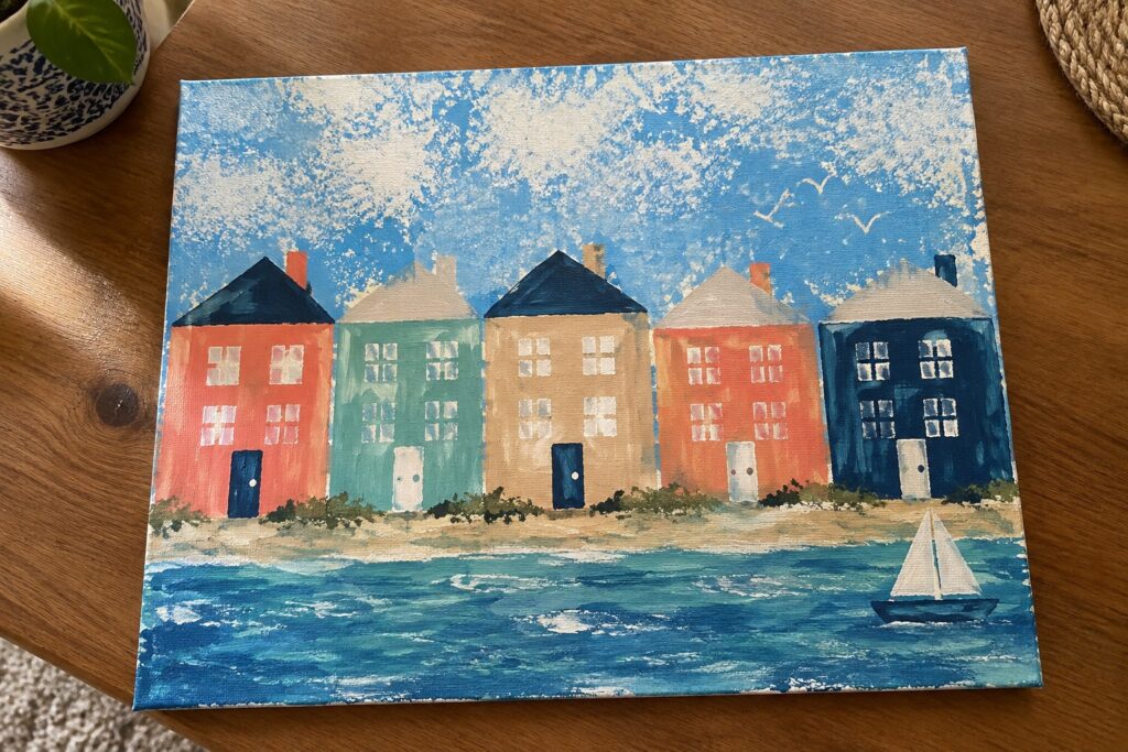 23 Easy Painting Ideas Using Simple Sponge and Stencil Techniques A canvas painting shows four brightly colored houses lined up beside teal water with three small sailboats and a yellow sun in the sky.