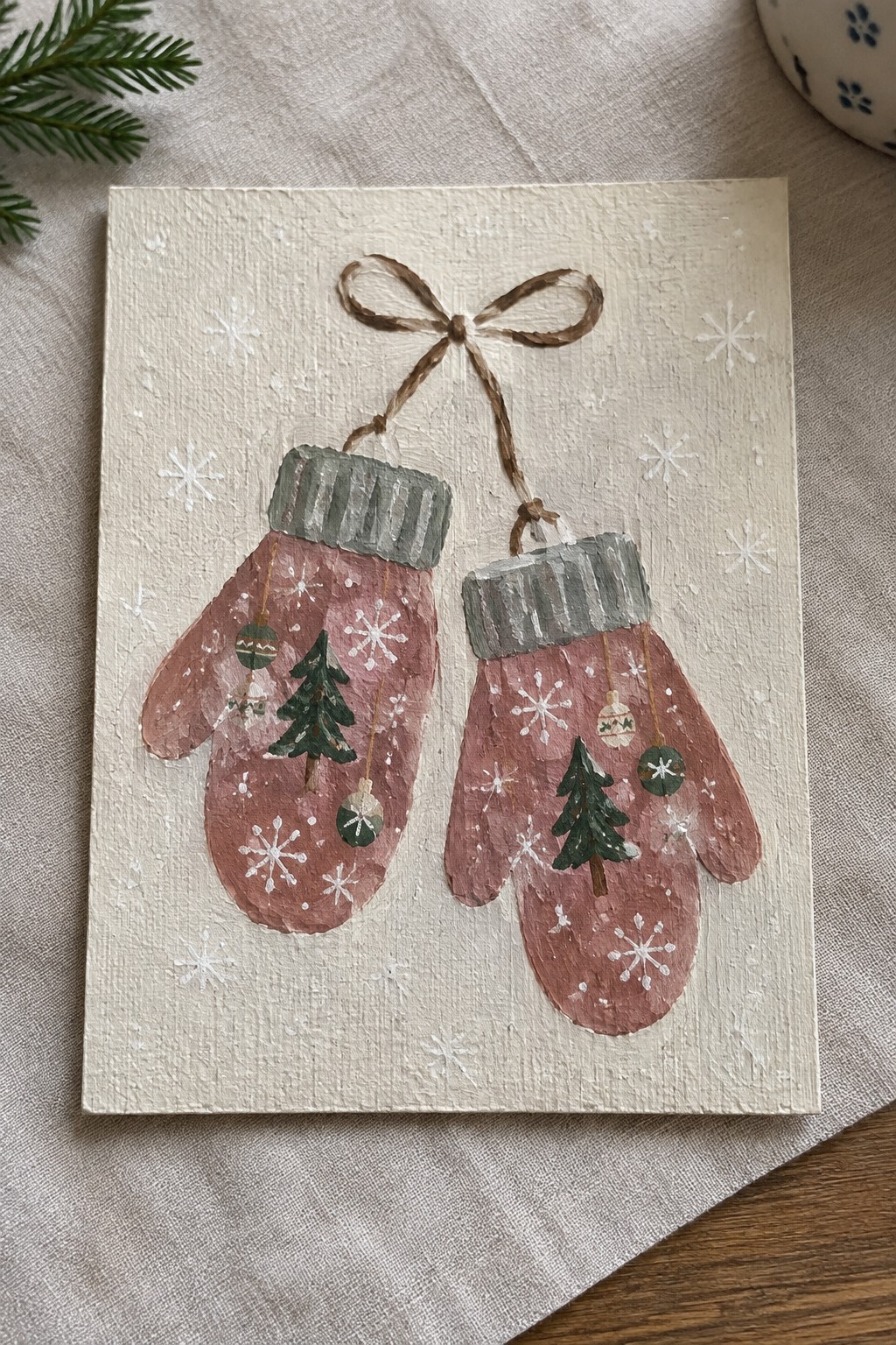 Two pink mittens with trees and snowflakes hanging from brown bow on canvas.