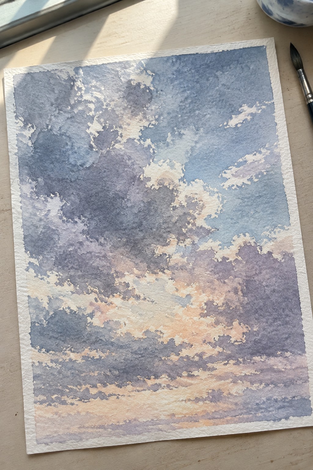 Watercolor sky painting with layered clouds in blue, purple, and orange tones on paper.
