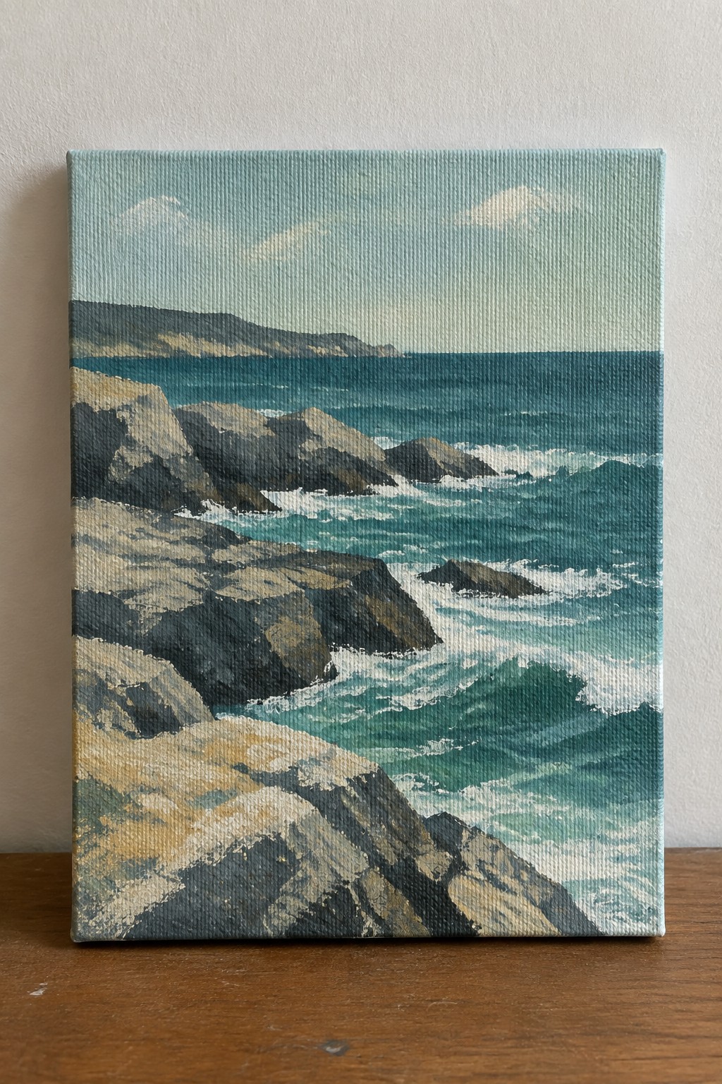 Seascape painting of rocky cliffs, turquoise waves, and distant hills on canvas.