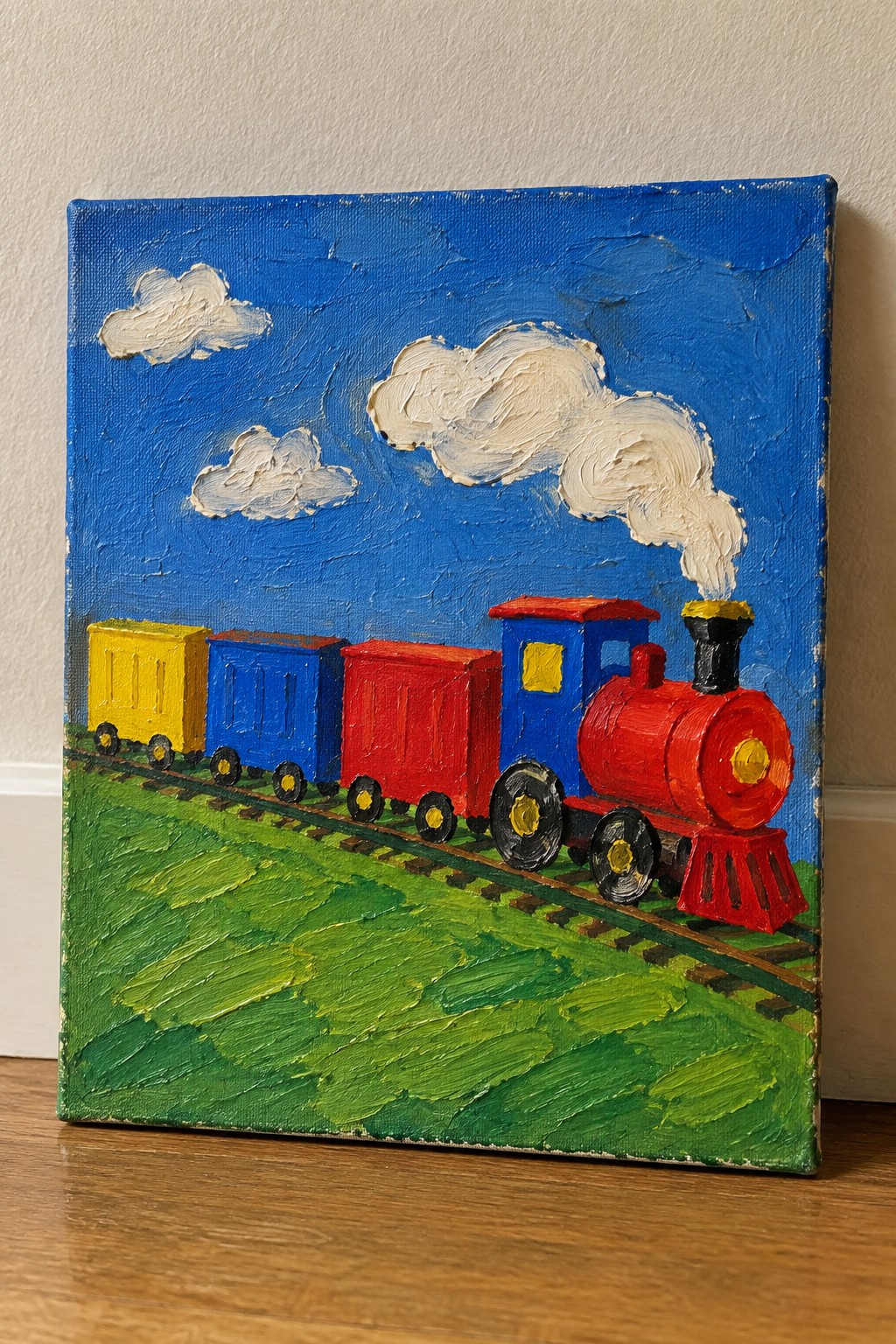 Thickly painted red toy train with colorful cars on tracks in green field under blue sky.