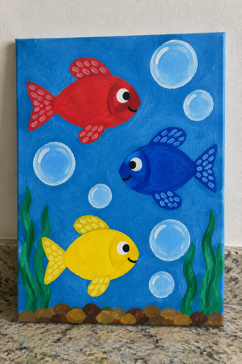 Three smiling fish in red, blue, and yellow swim among bubbles, seaweed, and pebbles.