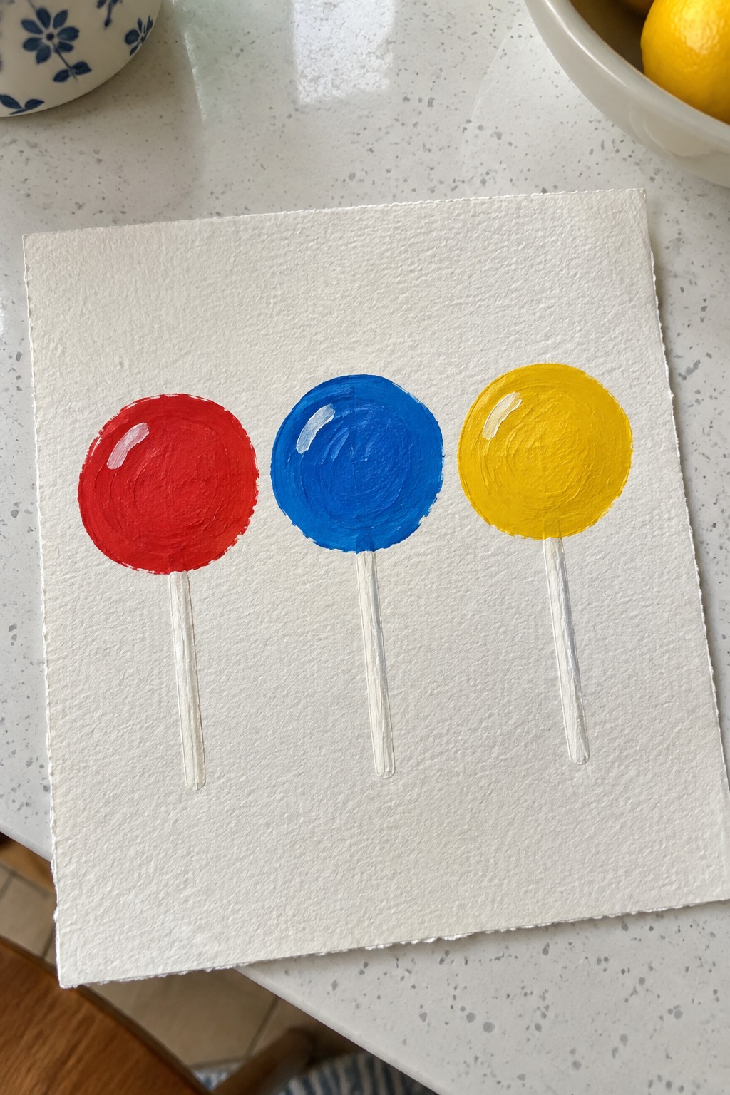 Three painted lollipops in red, blue, and yellow with white sticks on paper