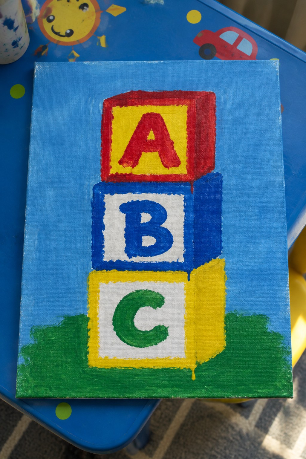 Colorful painting of stacked ABC blocks in red, blue, and yellow on blue canvas.