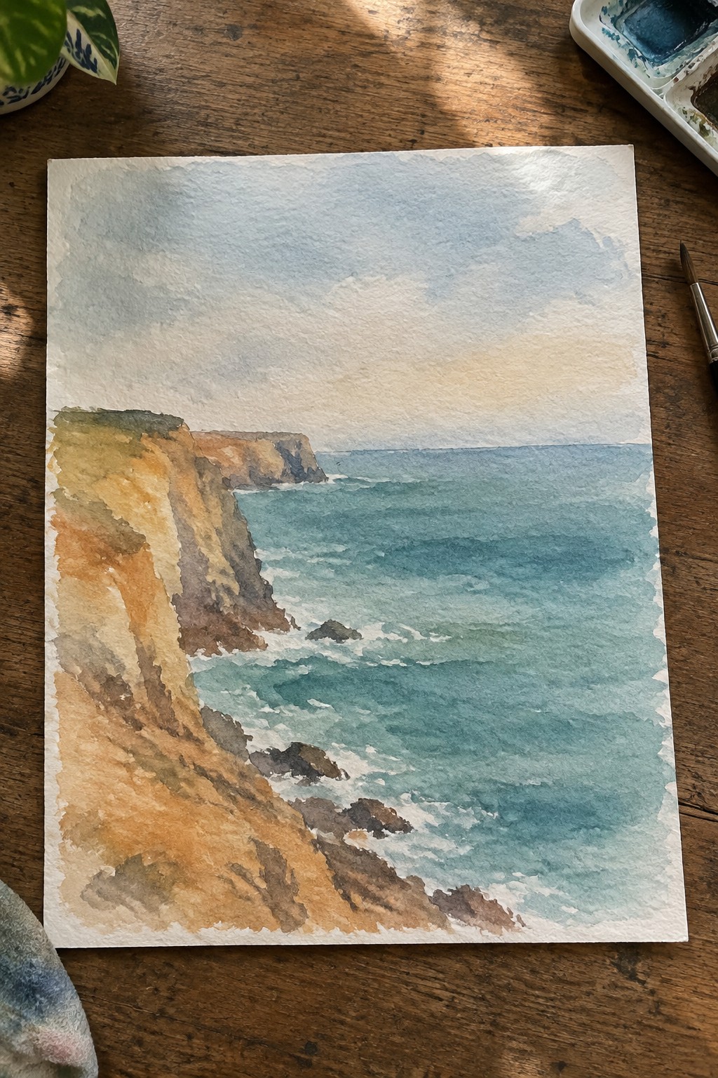 Watercolor seascape of cliffs and blue waves on wooden table with palette and brush