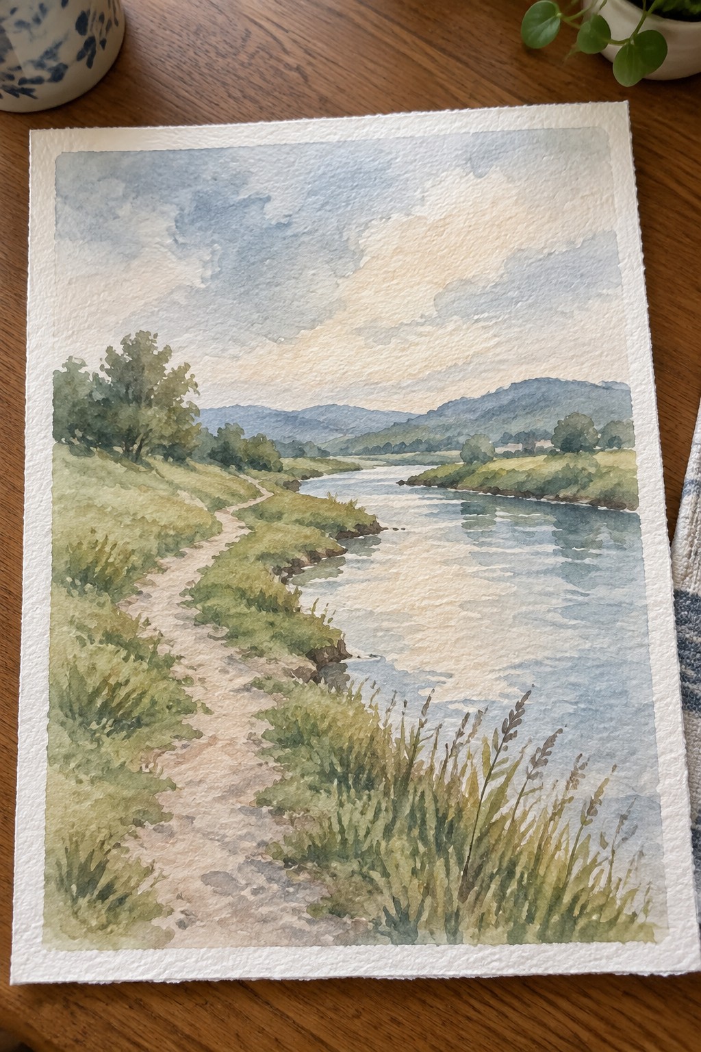 Watercolor landscape of winding river, dirt path, and green hills under cloudy sky