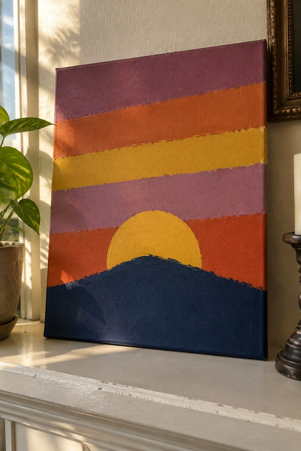 Striped sunset painting with yellow sun over blue mountain on windowsill