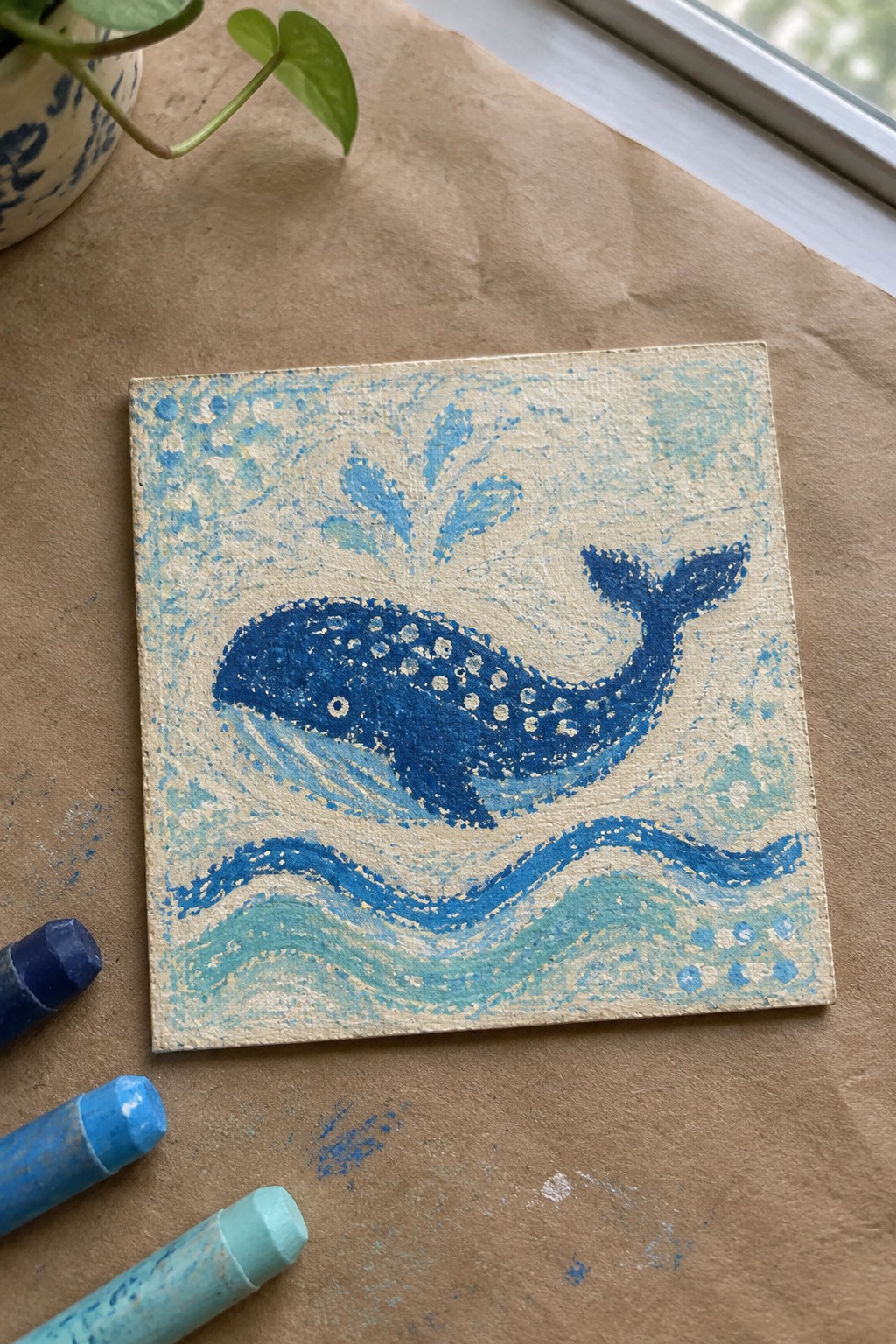 Blue whale painting with white spots over wavy blue-green water beside pastels