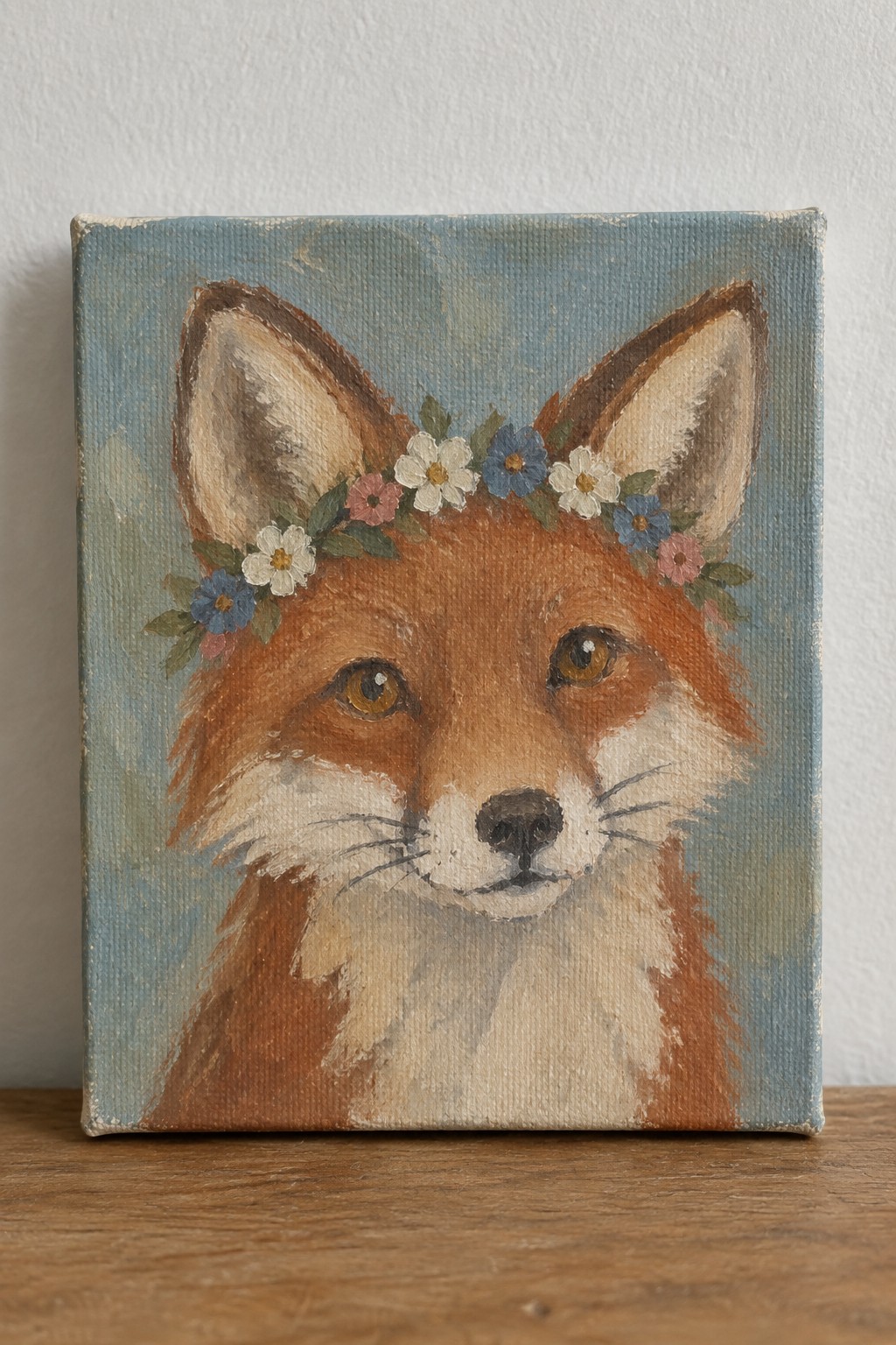 Fox with flower crown painted on canvas against blue background on wood.