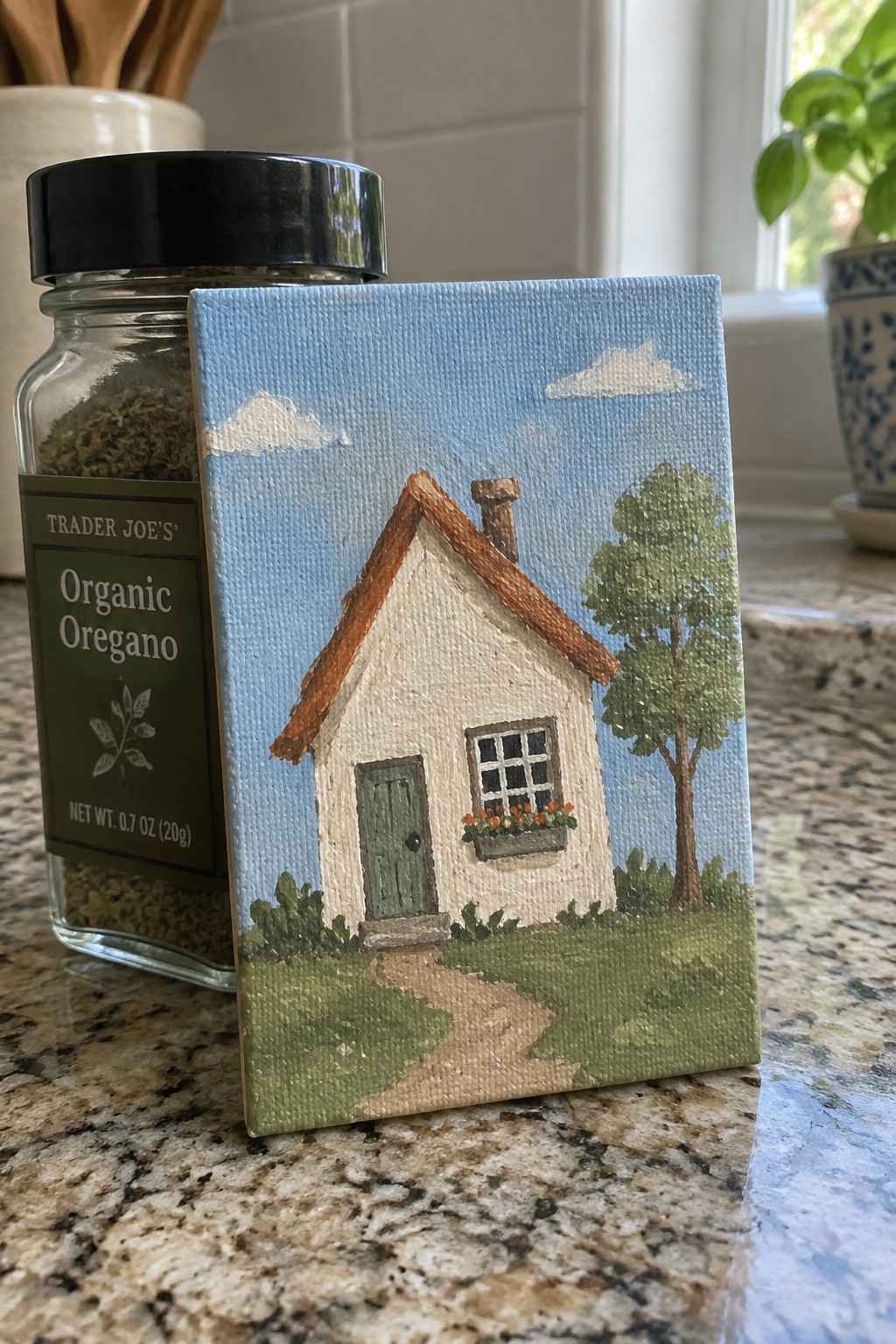 Small canvas painting of white cottage leaning against Trader Joe's oregano jar on counter
