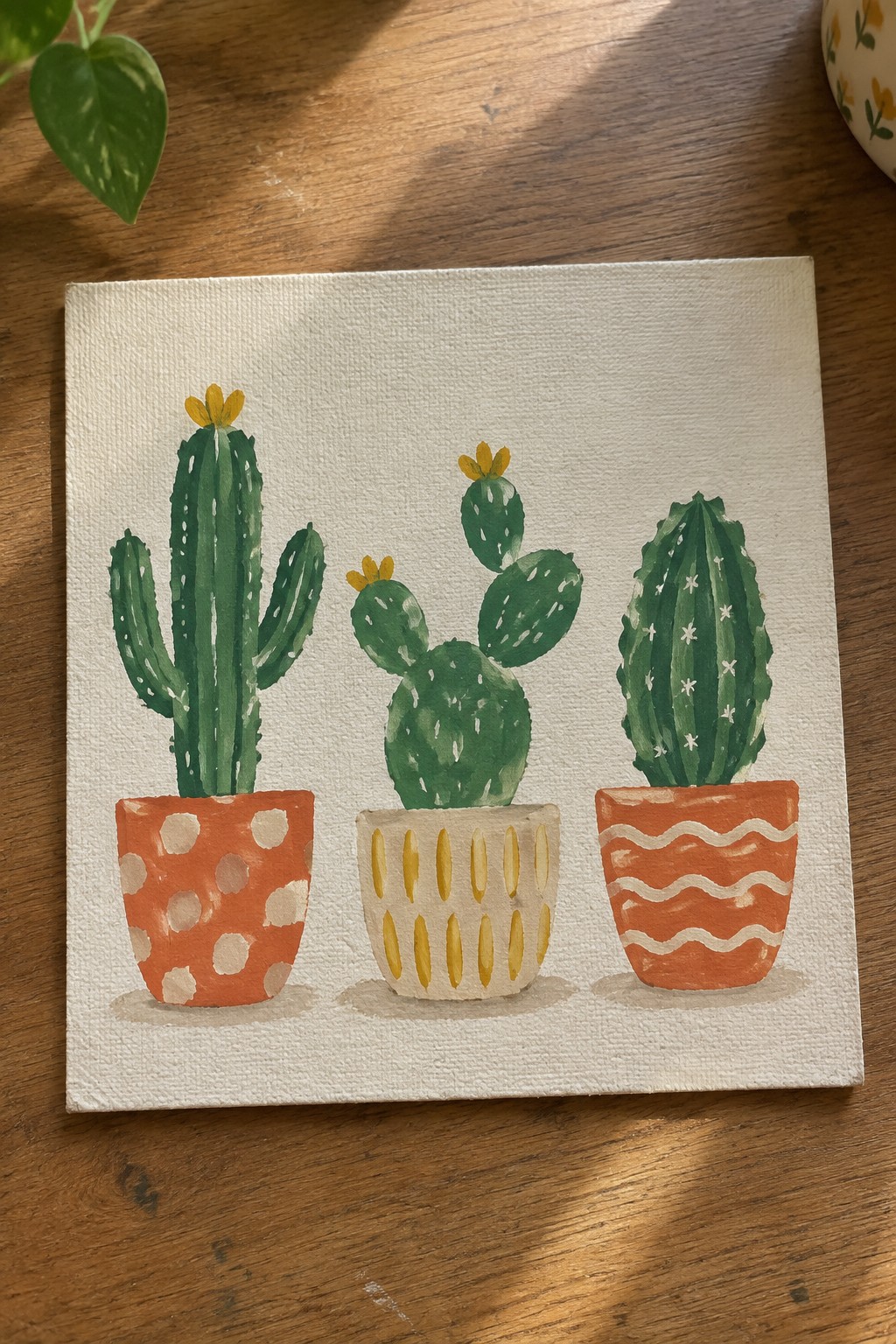 Three green cacti with yellow flowers painted in patterned orange and beige pots on a white canvas.