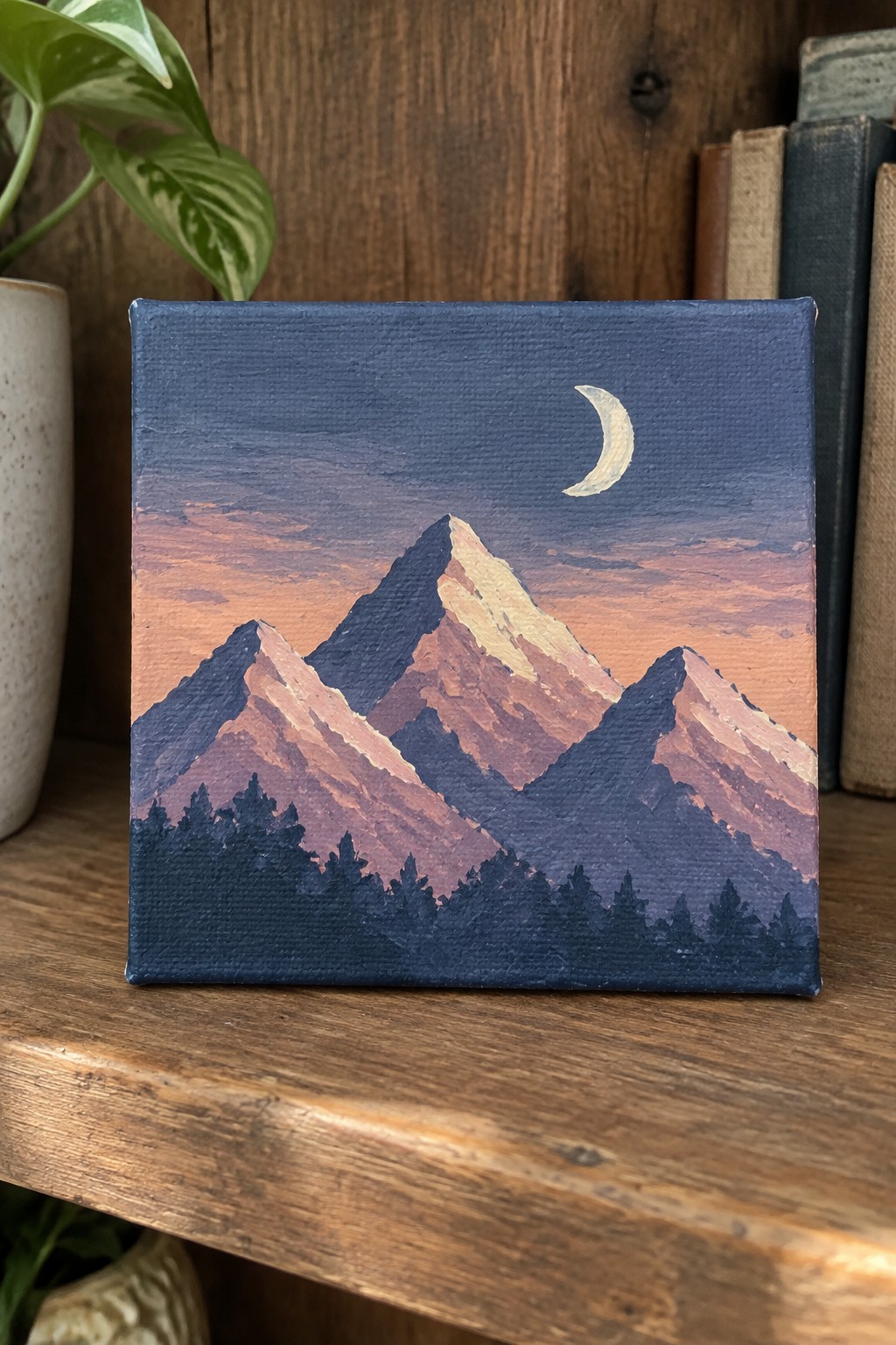 A small square canvas shows layered mountains in blue and orange tones with a crescent moon above and dark tree silhouettes at the base, sitting on a wooden shelf.