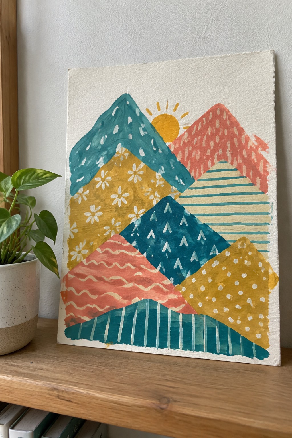 Colorful abstract mountain painting with geometric patterns in teal, yellow, and coral beside plant