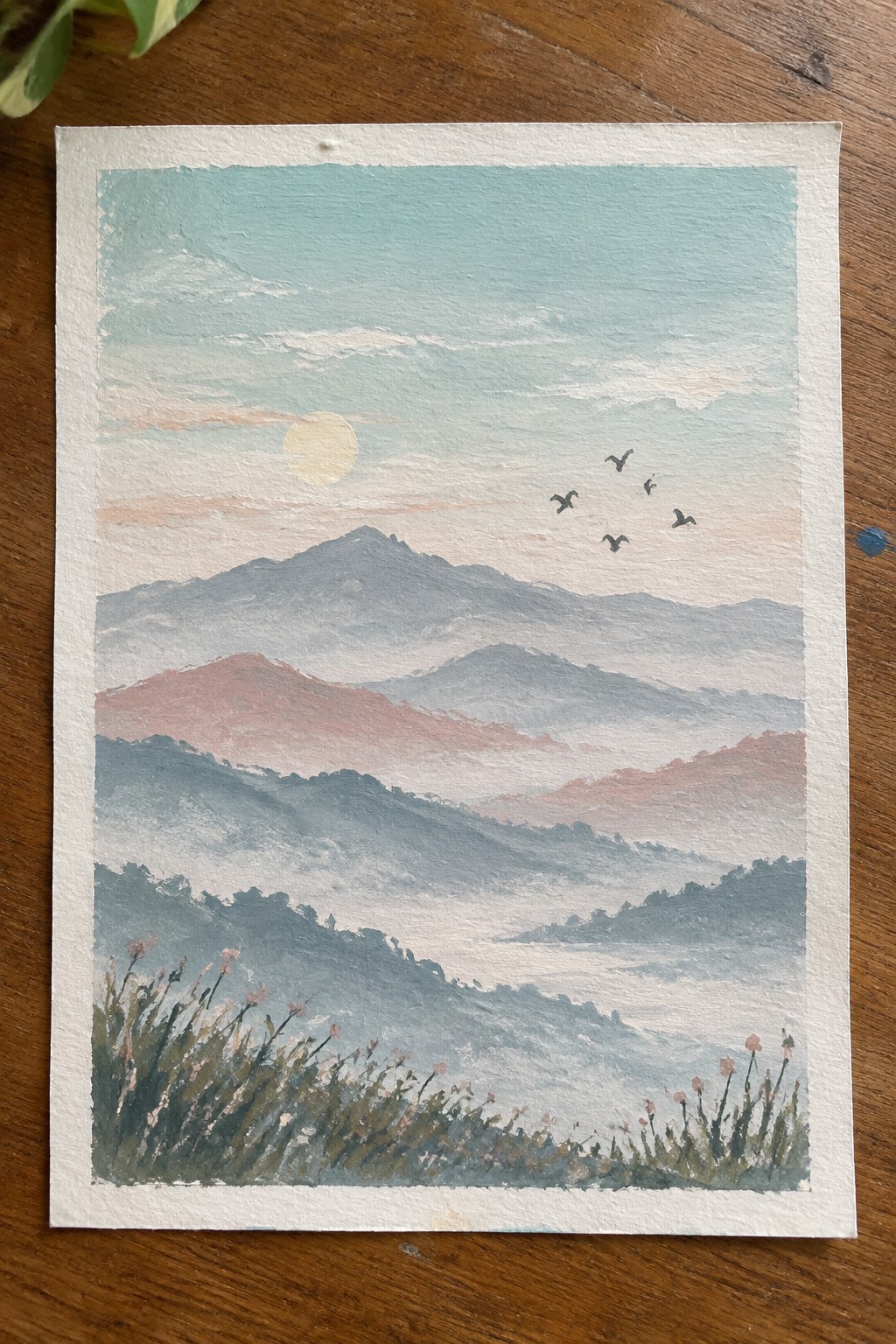 Watercolor landscape of misty mountains under pale sun with flying birds and foreground wildflowers.