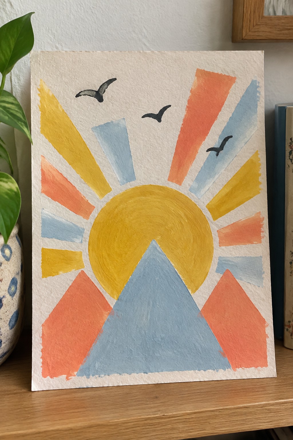 Watercolor painting of yellow sun with colorful rays above blue mountain and black birds.