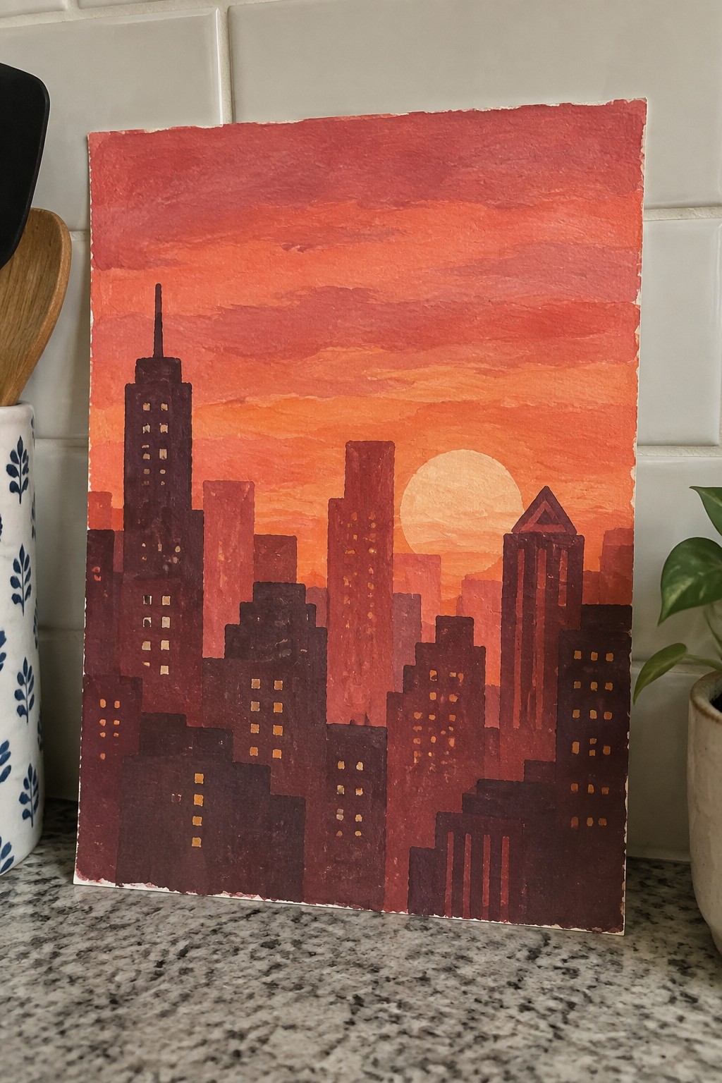 Watercolor city skyline painting with dark buildings and glowing windows at orange sunset.
