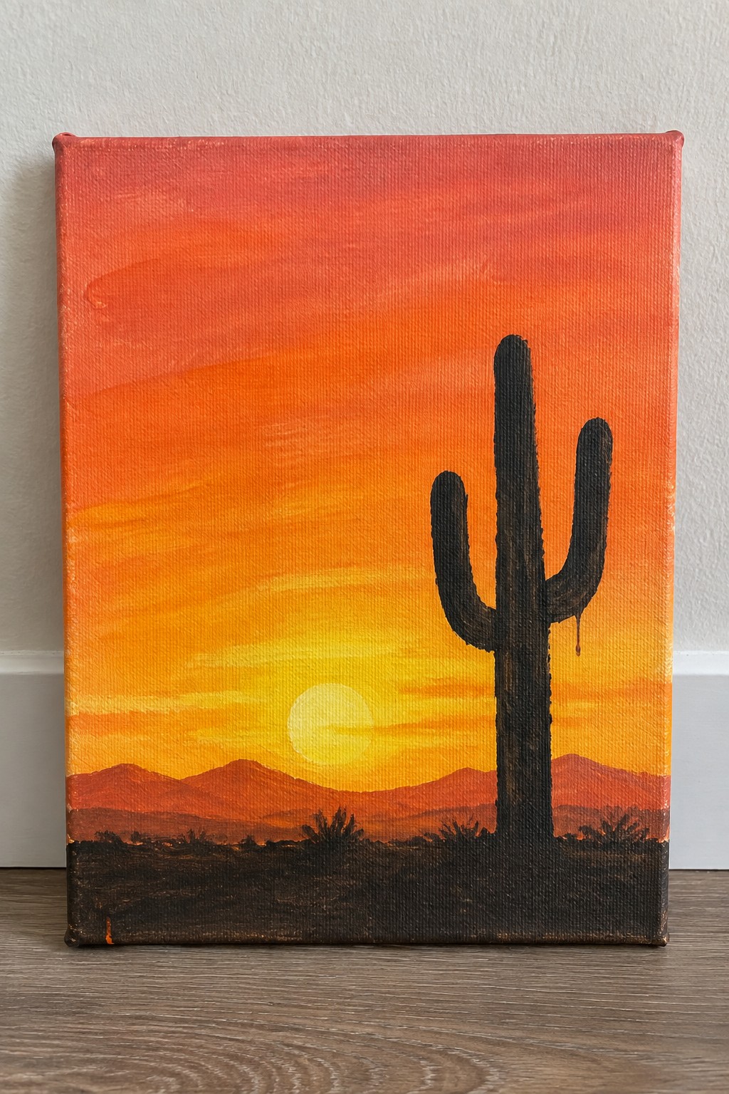 Vibrant orange-yellow sunset painting with silhouetted saguaro cactus and distant mountains.