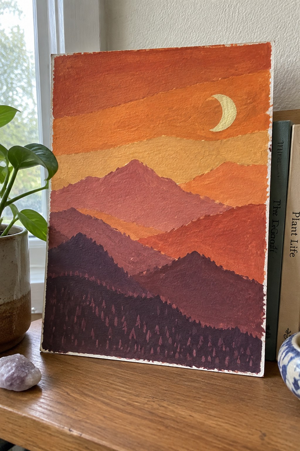 Layered red mountains under orange sunset sky with crescent moon and dark forest.